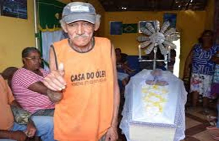 Homem dando joinha ao lado de um caixão num velório improvisado.